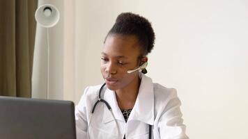 joven africano americano mujer médico con auriculares teniendo charla o consulta en ordenador portátil. hembra médico es vistiendo un auriculares, participación un médico en línea conferencia video