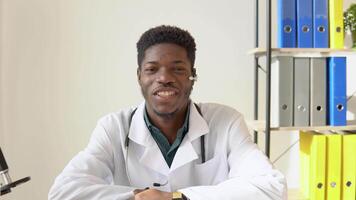 Young african american male doctor having chat or consultation on laptop looking directly to the camera. Confident young doctor holding seminar, webinar, lecture video