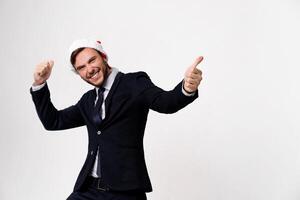 Young handsome caucasian guy in business suit and Santa hats stands on white background in studio smilie and showing thumbs up. photo