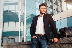 handsome young businessman with a beard and in a business suit standing on the street against the background of the office building next to a comfortable stylish leather bag. photo