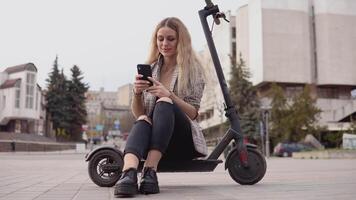 Young stylish blonde girl in a light jacket and jeans sits on an electric scooter and prints in a cell phone on the background of a quiet street video