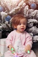 Small girl in the pink pajama sitting near the christmas tree playing with the garland photo