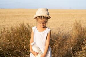 pequeño niña en pie trigo campo participación botella de Leche en mano vestido blanco vestir y Paja sombrero foto