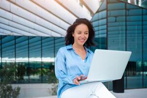 Smiling young businesswoman using laptop while sitting outside office building photo