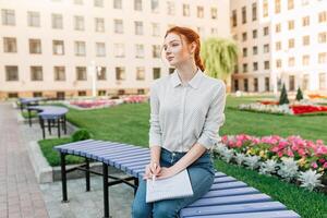 A beautiful young redhaired girl writes a romantic love letter sitting on a bench near the institute. Student love. Or homework photo