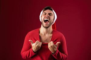 Young handsome caucasian guy in red sweater and Santa hats stands on red background in studio  Shows how hot he is photo