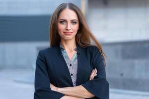 negocio mujer con largo pelo vestido negro chaqueta en pie al aire libre cerca corporativo oficina edificio manos doblada foto