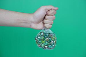 Close up hand hold net bag of marbles balls, small spherical object often made from glass. Green background. Concept. object use as toy for playing games or use for DIY craft decoration. photo