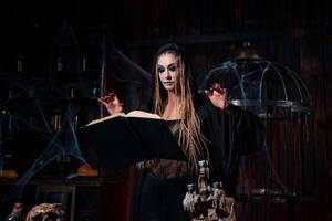 Halloween concept. Witch dressed black hood with dreadlocks standing dark dungeon room use magic book for conjuring magic spell photo