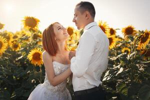 Bride in wedding white dresses and groom in whit shirt and tie standing hugging in the field of sunflowers photo
