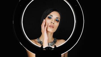 A beautiful young Caucasian girl with a beautiful make-up looks out from behind a circular lamp for portrait photo shooting