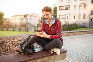 A redhair girl with freckles is dressed in a red checked shirt is sitting in a lotus pose in the park listening to music in wireless large headphones and texting message in the phone. Modern student photo
