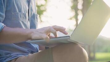 cerca arriba Disparo de masculino estudiante sentado abajo utilizando ordenador portátil computadora a el al aire libre trabajando parque, relajante trabajando ambiente, dorado hora atardecer, digital nómada laboral, mecanografía teclado escritura habilidades video