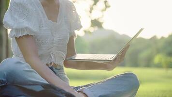 schließen oben ein Hand von jung Teenager halten Laptop Tippen auf ein Klaviatur, lesen auf Computer Bildschirm während Sitzung auf Grün Gras gegen Sonnenuntergang Fackeln auf sonnig Tag. Digital Hybrid Fernbedienung Arbeiten video