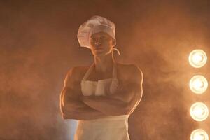 retrato de muscular cocinero vistiendo blanco delantal y cocinero sombrero, posando con doblada brazos en ahumado antecedentes foto