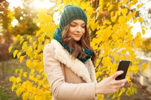 A beautiful young girl walks through the autumn park on the background of brightly colored leaves. Looks at your smartphone. Prints a message on the phone photo