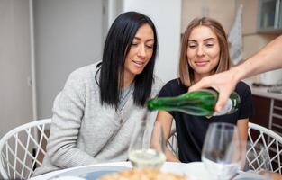 Friends meeting with wine and cake in the modern style kitchen. Young women smile and joke. A male hand in the foreground pours wine into glasses photo