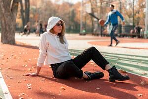 Outdoor portrait of young beautiful woman with long in sunglasses and a white hooded sweater sitting on the sportsground track photo