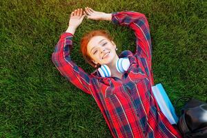 Beautiful young red-haired student girl with freckles lying on her back on grass and lawn view from above. Dressed in a red checkered shirt, resting after classes photo