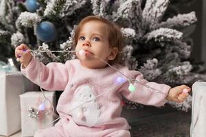 Small girl in the pink pajama sitting near the christmas tree playing with the garland photo