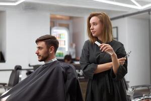 Handsome blue eyed man sitting in barber shop. Hairstylist Hairdresser Woman cutting his hair. Female barber. photo