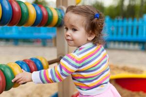 un pequeño niña con dos cruz es vestido en un a rayas vistoso chaqueta es jugando en el salvadera en el patio de recreo foto