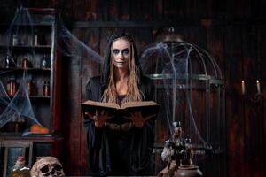 Halloween concept. Witch dressed black hood with dreadlocks standing dark dungeon room use magic book for conjuring magic spell photo