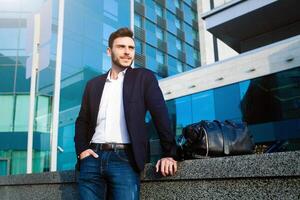 handsome young businessman with a beard and in a business suit standing on the street against the background of the office building next to a comfortable stylish leather bag. photo