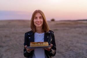 joven hermosa caucásico niña participación pastel en su manos en pie en naturaleza hermosa puesta de sol en antecedentes foto