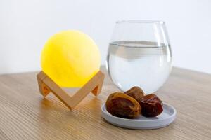 A small plate of dates fruit on a wooden table with a glass of water and lamp photo