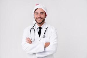 Young handsome doctor in white uniforme and Santa Claus hat standing in studio on white background loking at camera abd teeth smiling photo