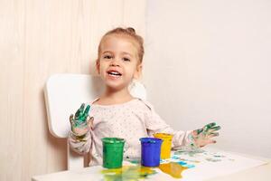 hermosa pequeño niña sorteos con dedo pinturas en un blanco sábana de papel. creativo niño desarrollo en jardín de infancia o gratis hora a hogar foto