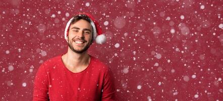 joven hermoso caucásico chico en rojo suéter y Papa Noel sombreros soportes en rojo antecedentes en estudio y y dientes sonriente foto