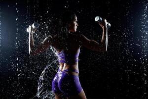 hermosa joven niña en púrpura ropa de deporte poses con pesas en agua estudio. gotas de agua untado acerca de su aptitud cuerpo. el Perfecto figura en el antecedentes de agua salpicaduras foto