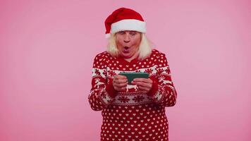 preocupado abuela en Navidad suéter con entusiasmo jugando carreras vídeo juegos en móvil teléfono video