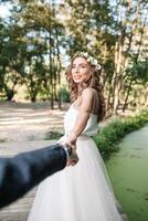 seguir yo mi amor concepto. atractivo joven mujer vestido en blanco Boda vestir participación mano de su novio y caminando en el hermosa jardín a contento futuro. foto