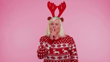 Senior old woman in Christmas sweater presses index finger to lips makes silence gesture sign secret video