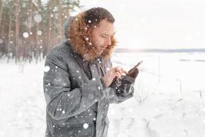 un joven hermoso hombre usos un móvil teléfono en un Nevado bosque. móvil redes adiccion. llamada para ayuda. llamada rescate foto