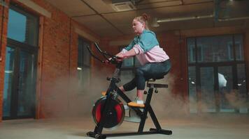 Athletic girl performing aerobic riding training exercises on cycling stationary bike in foggy gym video