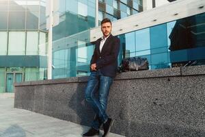 hermoso joven empresario con un barba y en un negocio traje en pie en el calle en contra el antecedentes de el oficina edificio siguiente a un cómodo elegante cuero bolsa. foto