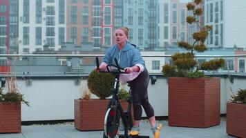 Healthy Caucasian woman exercising workout on stationary cycling machine bike on house rooftop gym video