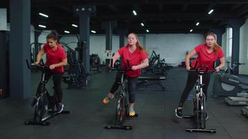 grupo de sonriente amigos mujer clase hacer ejercicio, capacitación, hilado en estacionario bicicleta a moderno gimnasio video