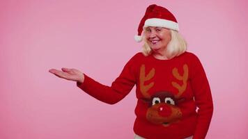 Grandmother in Santa Christmas sweater showing thumbs up and pointing empty place, advertising area video