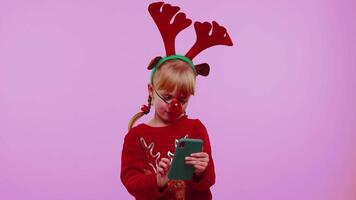 litet barn flicka i jul hatt använder sig av mobil telefon skriver ny posta på webb, handla uppkopplad, bläddring video