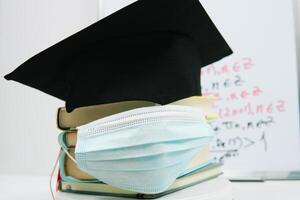 The black square hat of the master lies on the textbooks stacked in a column and decorated with a protective medical mask. Education concept photo