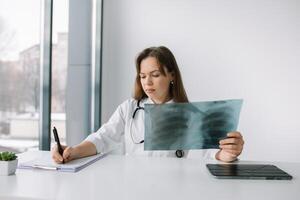 Young caucasian woman doctor wearing white medical coat and stethoscope holds the x-ray and makes notes in the hospital. Diagnosis of pneumonia photo