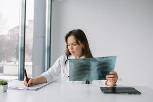 Young caucasian woman doctor wearing white medical coat and stethoscope holds the x-ray and makes notes. Diagnosis of tuberculosis photo