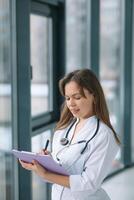 joven mujer médico vistiendo blanco médico Saco y estetoscopio escribe un médico historia en un cuaderno cerca el ventana foto