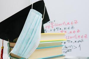 The black square hat of the master lies on the textbooks stacked in a column and decorated with a protective medical mask. Education concept photo