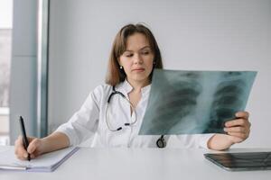 Young caucasian woman doctor wearing white medical coat and stethoscope holds the x-ray and makes notes in the hospital photo